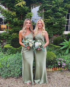 two beautiful women standing next to each other in front of some bushes and flowers on the ground