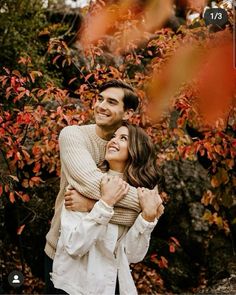 a man and woman hugging each other in front of trees with red leaves on them