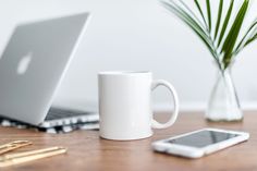 a white coffee mug sitting on top of a wooden table next to a laptop computer