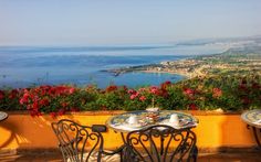 an outdoor table with two plates and cups on it overlooking the ocean in front of some flowers