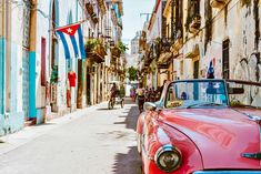 an old pink car parked on the side of a street next to tall buildings with flags hanging from them