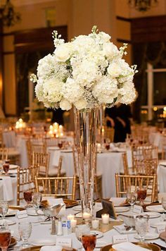 a tall vase filled with white flowers sitting on top of a table covered in gold chairs