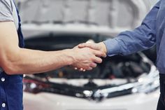 two men shaking hands in front of a car with the hood up and engine visible