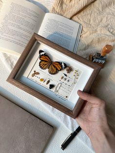 a person is holding a book and looking at a butterfly in a frame on a bed