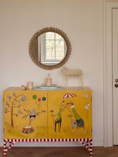 a yellow cabinet with paintings on it and a mirror hanging above the door, in front of a white wall