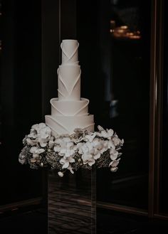 a white wedding cake sitting on top of a glass stand with flowers around it and greenery