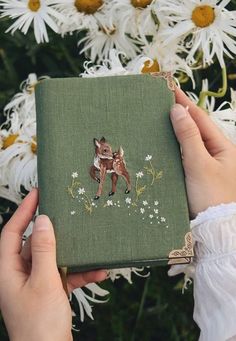 a person holding a small book in front of some flowers and daisies, with the image of a fox on it's cover