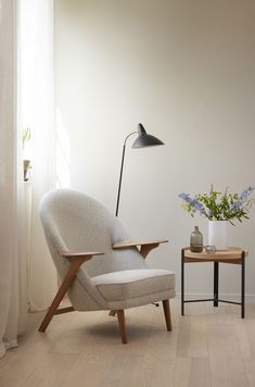 a white chair sitting in front of a window next to a table with a vase on it