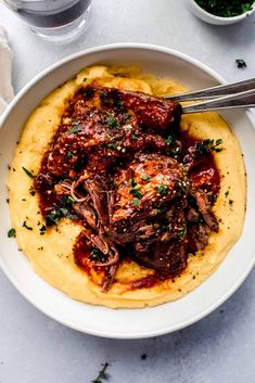 a white bowl filled with meat and sauce on top of mashed potatoes, garnished with parsley