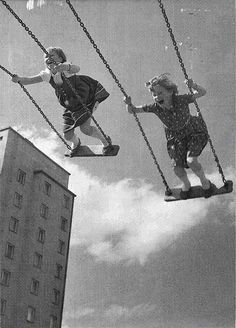 two children on swings in the air with buildings in the back ground and clouds in the sky