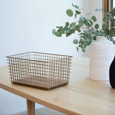 a basket sitting on top of a wooden table next to a vase with green leaves