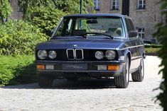 an old bmw is parked in front of some bushes and trees on a cobblestone road