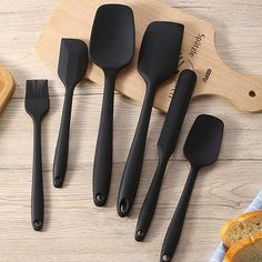 five black kitchen utensils on a cutting board with bread and knifes next to it