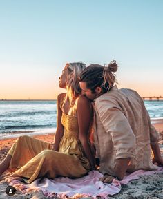 two women sitting on the beach with their arms around each other as they look at the water