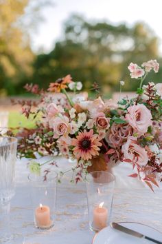 an arrangement of flowers and candles on a white table cloth with silverware in the foreground