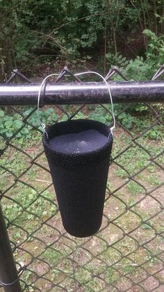 a black bird feeder hanging from a chain link fence next to some bushes and trees