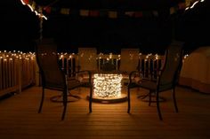 some chairs are sitting on a deck with lights in the dark and there is a fire pit surrounded by string lights