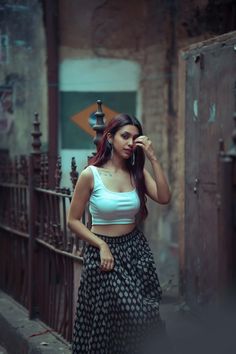 a woman in a white top and black skirt is standing on the street with her hand to her face