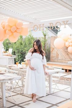 a pregnant woman wearing a white dress standing in front of an outdoor table with balloons