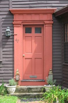 a red door is on the side of a gray house with potted plants in front