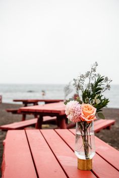 a vase with flowers sitting on top of a wooden table near the ocean and benches