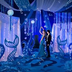 a man and woman standing in front of a stage decorated with blue curtains, lights and decorations