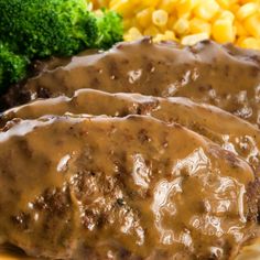 two pieces of meat covered in gravy next to broccoli and corn