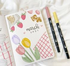 an open planner book next to two markers and pencils on a white bed sheet