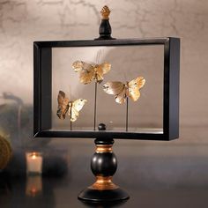 three golden butterflies in a glass frame on a black table next to a candle and vase