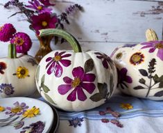 three white pumpkins with purple flowers painted on them, sitting next to two plates