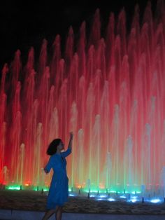 a woman standing in front of a colorful fountain at night with her hand up to the sky