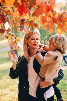 a woman holding a child in her arms under a tree with autumn leaves on it