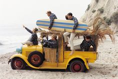 a group of men riding on the back of a yellow truck down a sandy beach