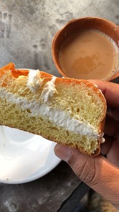 a person holding a piece of bread with cream on it next to a cup of coffee