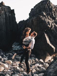 a man carrying a woman on his back while standing on rocks near the ocean with mountains in the background