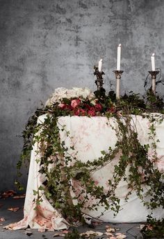 an arrangement of flowers and candles on a table with cloth draped over it in front of a concrete wall