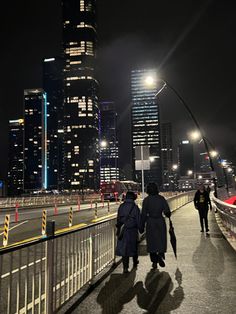 two people walking across a bridge in the city at night with tall buildings behind them