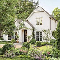 a white house with lots of bushes and flowers around it's front door, surrounded by greenery