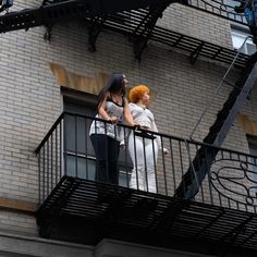 two women are standing on the balcony of a brick building and talking to each other