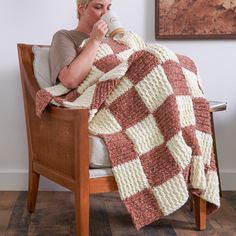 a woman sitting on a chair drinking from a coffee cup while wrapped in a crocheted blanket