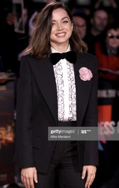 a woman in a tuxedo and bow tie smiles as she stands on the red carpet