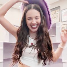 a woman with long brown hair wearing a purple hat and white tank top smiling at the camera