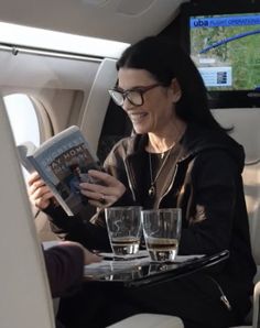 a woman is sitting in an airplane reading a book and drinking some wine while another person looks on