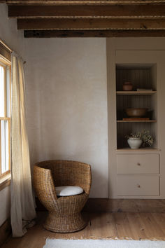 a wicker chair sitting in front of a window next to a wooden shelf with bowls on it