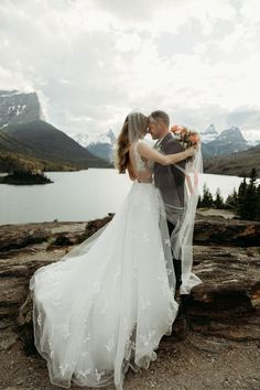 a bride and groom standing on top of a mountain next to a lake with mountains in the background