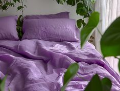 a bed covered in purple sheets and pillows next to a potted plant with green leaves