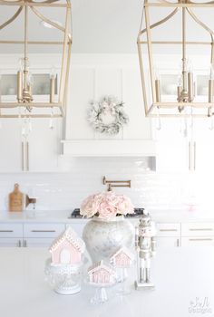 a white kitchen with gold accents and pink flowers on the countertop in front of two chandeliers