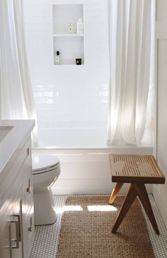 a white bathroom with a wooden bench in front of the toilet and shower curtained window