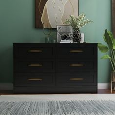 a black dresser sitting in front of a painting on the wall next to a potted plant