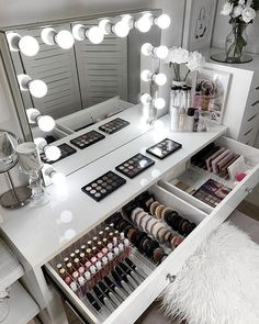 a vanity with lights and makeup on it in a room next to a mirror that has drawers full of cosmetics